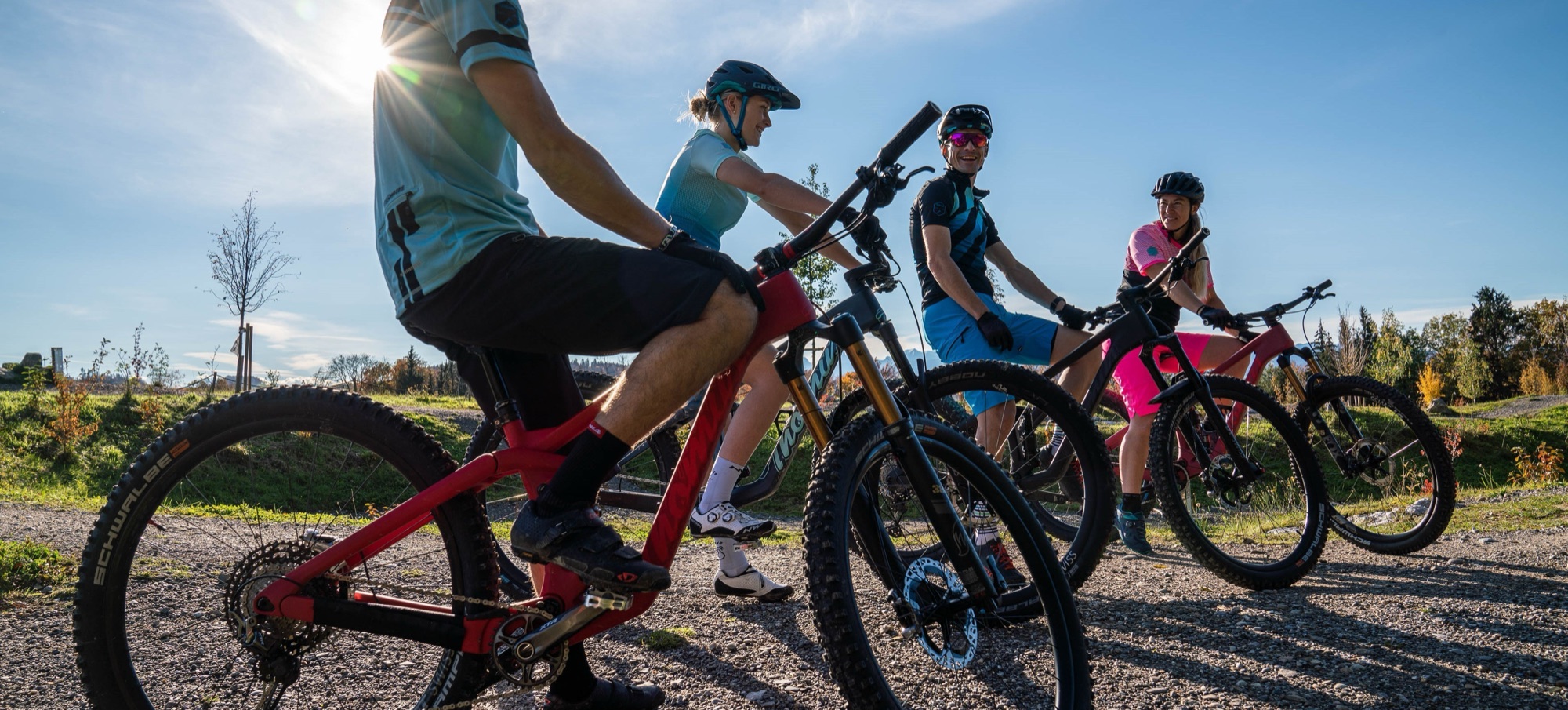 Vier Biker:innen stehen auf einem Schotterhügel, die Sonne strahlt zwischen ihnen hindurch